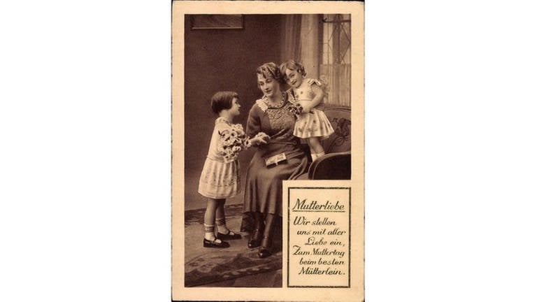 Mutter mit ihren zwei kleinen Töchtern, die ihr Blumen schenken (Foto): Glückwunschkarte zum Muttertag, um 1933, mit der Aufschrift Mutterliebe – Wir stellen uns mit aller Liebe ein, zum Muttertag beim besten Mütterlein